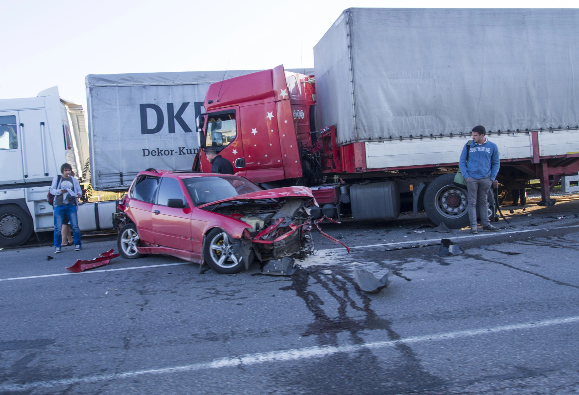 Damaged red car wedged beneath two semi trucks after a roadway crash
